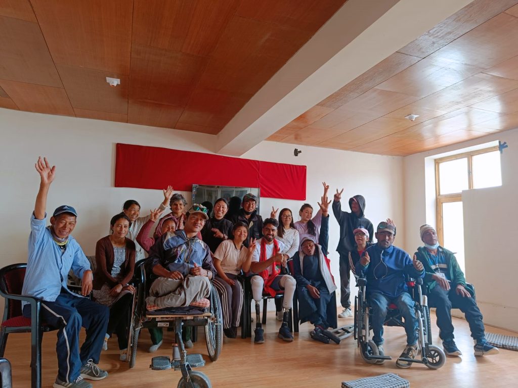 A group of wheelchair and non-wheelchair users are seen in a room, alongwith Tinkesh, posing with their hands raised in the air, cheering, and seeming happy.