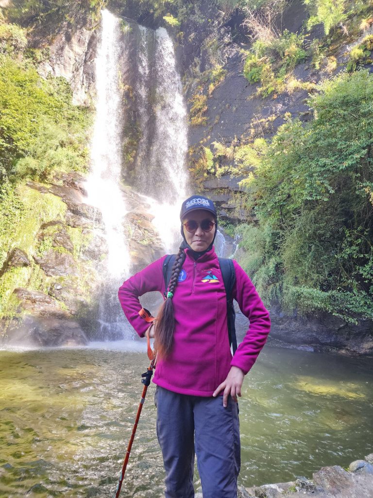 a blind woman with a cane wearing a pink jacket, a cap, and sunglasses standing in front of a waterfall
