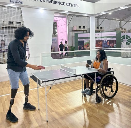 Tinkesh is seen happily playing table tennis with an opponent using a wheelchair in a brightly lit room with white walls and large glass windows.