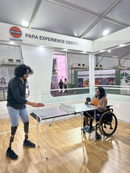 Tinkesh is seen happily playing table tennis with an opponent using a wheelchair in a brightly lit room with white walls and large glass windows.