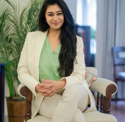 Dr. Manasi Shankar in a grey pant suit, with a green shirt. long black hair, seated on a wood chair with beige cushions