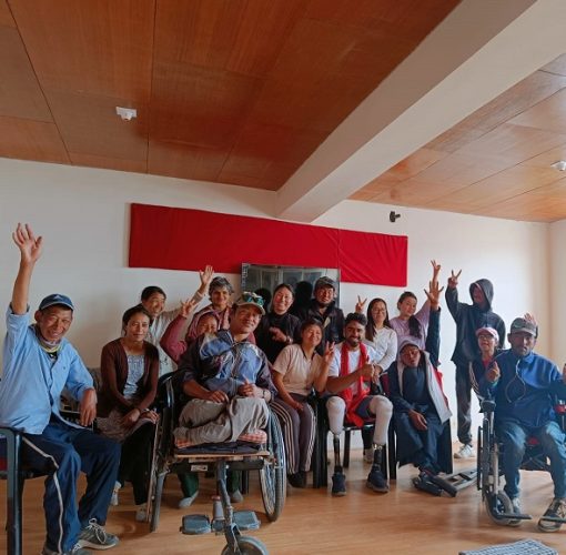 A group of wheelchair and non-wheelchair users are seen in a room, alongwith Tinkesh, posing with their hands raised in the air, cheering, and seeming happy.